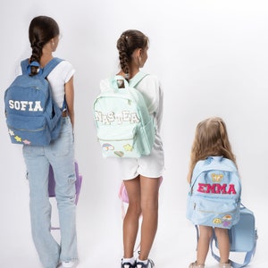 Op de afbeelding: Drie kinderen dragen rugzakken met hun namen Sofia, Nastia en Emma erop geborduurd. De rugzakken zijn blauw, lichtgroen en lichtblauw. De kinderen staan voor een witte achtergrond.