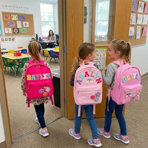 May include: Three young girls wearing pink backpacks with personalized names and colorful patches. The backpacks have the names "BAYLEE", "ADALYN", and "OLIVIA" on them. They are walking through a doorway into a classroom.