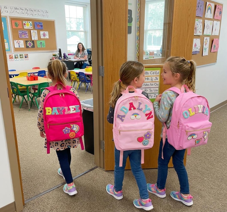 May include: Three young girls wearing pink backpacks with personalized names and colorful patches. The backpacks have the names "BAYLEE", "ADALYN", and "OLIVIA" on them. They are walking through a doorway into a classroom.