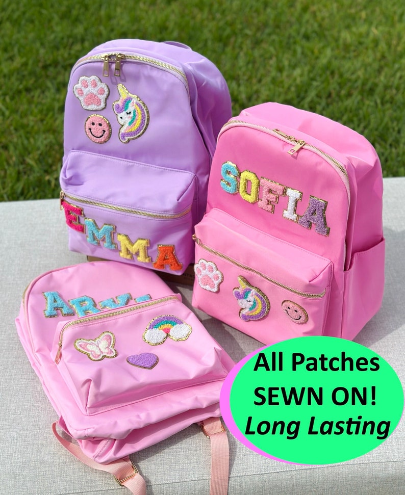 May include: Three colorful backpacks in pink and purple, adorned with embroidered patches and personalized names. Each backpack features a front pocket, gold zippers, and decorative patches including a paw print, unicorn, and smiley face. The text on the image says "All Patches SEWN ON! Long Lasting".