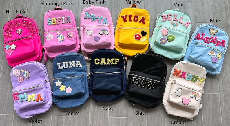 May include: A collection of personalized backpacks in various colors, including hot pink, flamingo pink, baby pink, yellow, mint, blue, violet, denim, navy, black, and cream. Each backpack features a name patch and decorative patches.