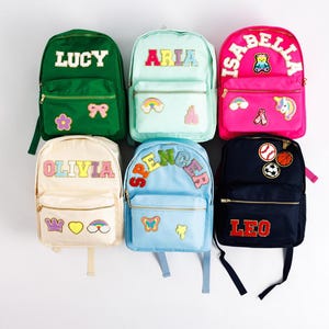 May include: Six colorful backpacks with personalized names and decorative patches. The backpacks are in various colors, including green, pink, blue, and cream. Each backpack has a name in bold letters, such as "LUCY" and "OLIVIA".