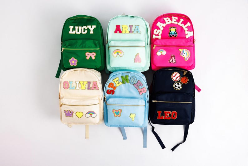 May include: Six colorful backpacks with personalized names and decorative patches. The backpacks are in various colors, including green, pink, blue, and cream. Each backpack has a name in bold letters, such as "LUCY" and "OLIVIA".