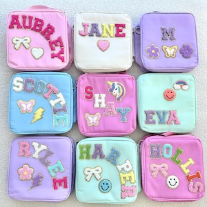 May include: Nine colorful lunch bags with personalized names in various colors. Each bag features a different name, such as "Aubrey," "Jane," and "M," with decorative patches like butterflies, flowers, and rainbows.