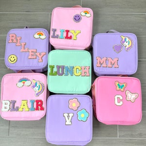 May include: Seven colorful lunch bags in shades of pink, purple, and green. Each bag features a name or word in sequined letters, along with decorative patches like rainbows, flowers, and a unicorn. The bags have a handle and a zipper.