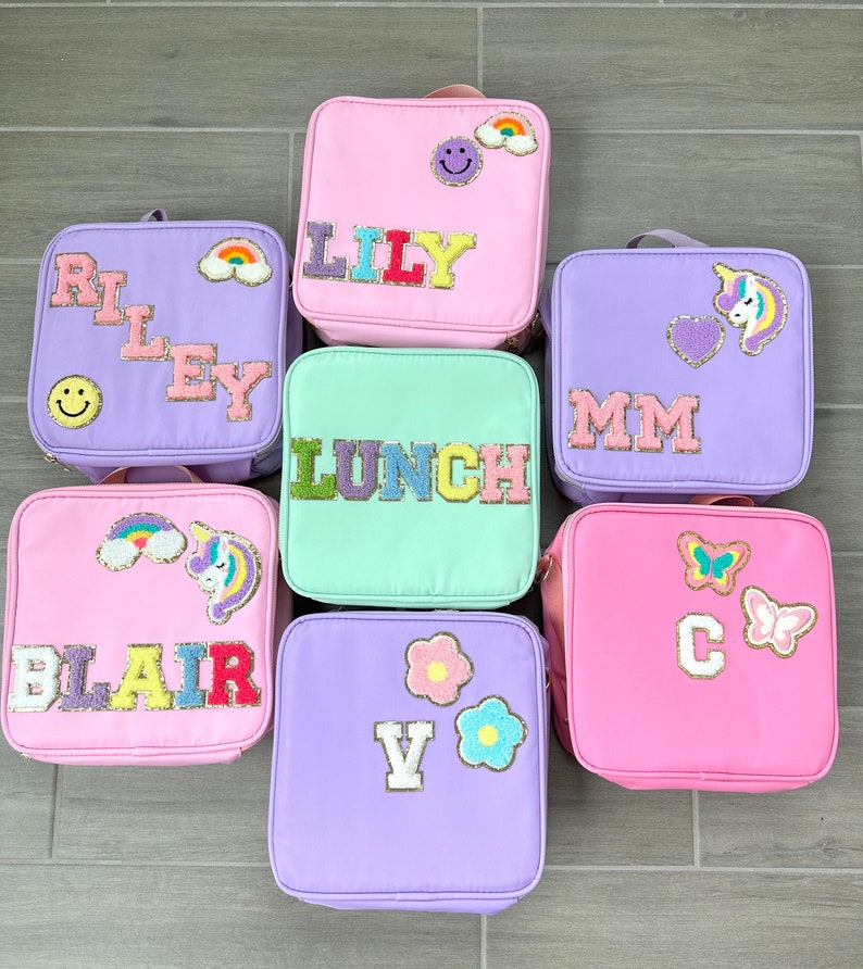 May include: Seven colorful lunchboxes in various pastel shades, including pink, purple, and mint green. Each lunchbox is adorned with decorative patches, such as rainbows, smiley faces, unicorns, butterflies, and personalized names or initials in glittery letters.
