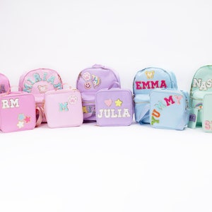 May include: Six pastel-colored backpacks and lunch bags with personalized names and colorful patches. The backpacks are pink, purple, light blue, and light green. The lunch bags are pink, purple, light blue, and light green. The names on the backpacks and lunch bags are "Sophia", "Miriam", "Julia", "Emma", "Nastea", "RM", "M", "Yummy", and "Snack".