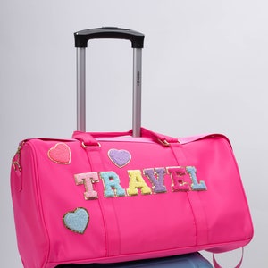以下が含まれることがあります： カラフルな文字で「TRAVEL」と書かれた、鮮やかなピンク色の旅行用ダッフルバッグ。ハート型のパッチとシルバーのハンドルが付いています。水色のキャリーケースの上に置かれています。