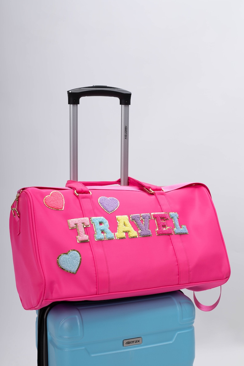以下が含まれることがあります： カラフルな文字で「TRAVEL」と書かれた、鮮やかなピンク色の旅行用ダッフルバッグ。ハート型のパッチとシルバーのハンドルが付いています。水色のキャリーケースの上に置かれています。