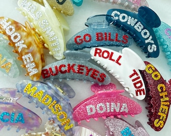May include: A collection of colorful, glittery hair clips with various names and phrases, including "Mizzou", "Go Fins", "Daniela", "Cowboys", "Go Bills", "Roll Tide", "Go Chiefs", "Buckeyes", "Madison", "Doina", "Hook Em", "Taylor", "Gigi", "Bride", and "CIA".