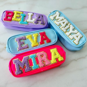 May include: Four personalized pencil cases in various colors: purple, light blue, and hot pink. Each case features a name in colorful, textured letters with gold trim. The names are "BELLA", "MAYA", "EVA", and "MIRA".
