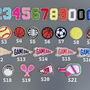 K&ouml;nnte beinhalten: Eine Sammlung von aufgestickten Aufn&auml;hern mit Zahlen, Sportb&auml;llen und Wimpeln. Die Aufn&auml;her umfassen die Zahlen 0-9, Fu&szlig;b&auml;lle, Baseb&auml;lle, Basketb&auml;lle, Tennisb&auml;lle, American Footballs und Wimpel mit "GAME DAY"-Text.