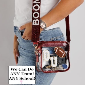 May include: A clear, red crossbody bag with a sequined strap that reads "BOOMER" in white letters. The bag contains a football patch, and the letters "O" and "U". The text "We Can Do ANY Team! ANY School!" is also visible.