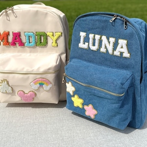 May include: Two personalized backpacks. One is cream-colored with the name "MADDY" in colorful, textured letters. The other is denim blue with "LUNA" in white, gold-trimmed letters. Both have front pockets and decorative patches.