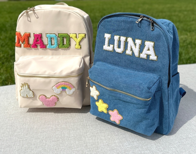 May include: Two personalized backpacks. One is cream-colored with the name "MADDY" in colorful, textured letters. The other is denim blue with "LUNA" in white, gold-trimmed letters. Both have front pockets and decorative patches.