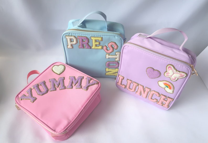 May include: Three colorful lunch bags in pastel shades of pink, blue, and lavender. Each bag features embroidered text and decorative patches. The pink bag says "YUMMY", the blue bag says "PRESTON", and the lavender bag says "LUNCH".