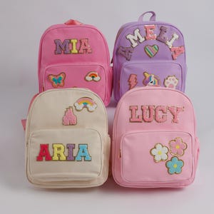 May include: Four colorful backpacks, each with a name embroidered on the front. The backpacks are pink, lavender, cream, and light pink. Each backpack has decorative patches, including butterflies, rainbows, and flowers.