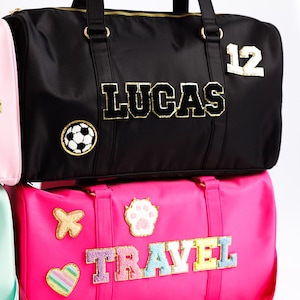 以下が含まれることがあります： ゴールドのアクセントが付いた黒いダッフルバッグ。「LUCAS」の金色の文字とサッカーボールのパッチ。下のピンクのダッフルバッグには、カラフルな文字で「TRAVEL」の文字と様々なパッチが付いています。