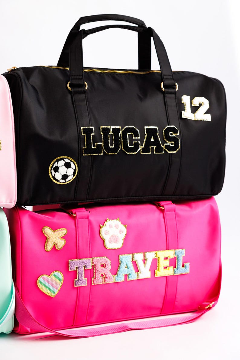 以下が含まれることがあります： ゴールドのアクセントが付いた黒いダッフルバッグ。「LUCAS」の金色の文字とサッカーボールのパッチ。下のピンクのダッフルバッグには、カラフルな文字で「TRAVEL」の文字と様々なパッチが付いています。
