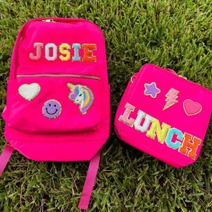 May include: A bright pink backpack and lunch bag set. The backpack has the name "JOSIE" in colorful embroidered letters, along with a heart, smiley face, and unicorn patch. The lunch bag says "LUNCH" and features star, lightning bolt, and heart patches.