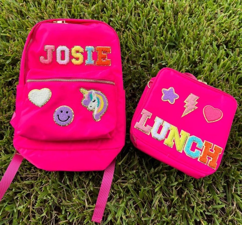 May include: A bright pink backpack and lunch bag set. The backpack has the name "JOSIE" in colorful embroidered letters, along with a heart, smiley face, and unicorn patch. The lunch bag says "LUNCH" and features star, lightning bolt, and heart patches.