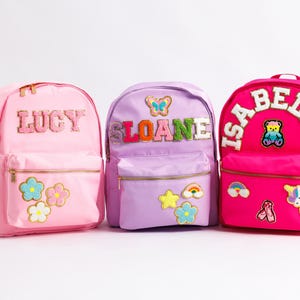 May include: Three colorful backpacks, pink, lavender, and fuchsia, each personalized with a name. The pink backpack says "LUCY" and has flower patches. The lavender backpack says "SLOANE" with a butterfly. The fuchsia backpack says "ISABELLA" with a teddy bear.