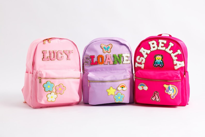 May include: Three colorful backpacks, pink, lavender, and fuchsia, each personalized with a name. The pink backpack says "LUCY" and has flower patches. The lavender backpack says "SLOANE" with a butterfly. The fuchsia backpack says "ISABELLA" with a teddy bear.