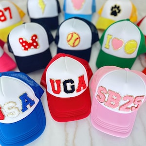 May include: A collection of trucker hats with white fronts and mesh backs in various colors. Designs include a lightning bolt, heart, paw print, Minnie Mouse bow, baseball, and text. The hats feature the text "UGA", "USA", and "SB25".