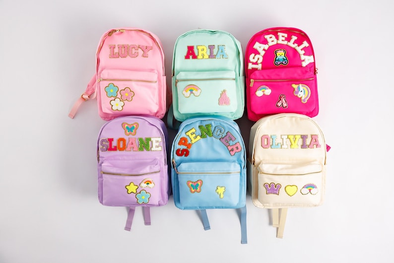 May include: Six colorful backpacks with personalized names. The backpacks are in pink, mint green, hot pink, lavender, light blue, and cream. Each backpack has a name patch and decorative patches like flowers, rainbows, and butterflies.