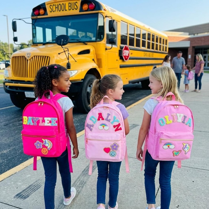 May include: Three young girls wearing pink backpacks with personalized names and colorful patches, walking towards a yellow school bus. The backpacks have names like "BAYLEE", "ADALYN", and "OLIVIA". The scene is set in front of a school building.