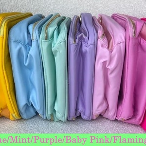 May include: A collection of colorful, rectangular pouches in various shades, including black, yellow, blue, mint, purple, baby pink, flamingo pink, and hot pink. Each pouch has a gold zipper and a matching interior lining.