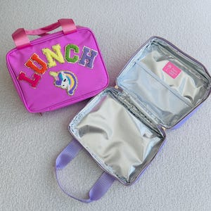 Op de afbeelding: Een roze lunchtas met het woord "LUNCH" in kleurrijke geborduurde letters en een eenhoornpatch. De tas heeft een handvat aan de bovenkant en een ritssluiting. De binnenkant is ge&iuml;soleerd met een zilveren voering.