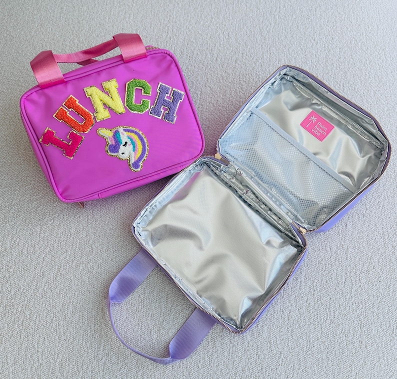Op de afbeelding: Een roze lunchtas met het woord "LUNCH" in kleurrijke geborduurde letters en een eenhoornpatch. De tas heeft een handvat aan de bovenkant en een ritssluiting. De binnenkant is ge&iuml;soleerd met een zilveren voering.
