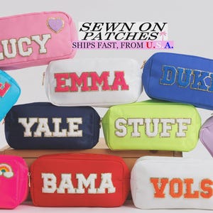 May include: A collection of colorful cosmetic bags with embroidered text and patches. Bags are in various colors including pink, blue, white, and red. Text includes names and words like "LUCY", "EMMA", "DUKE", "DANCE", "YALE", "STUFF", "RB", "BAMA", "VOLS", and "LSU".