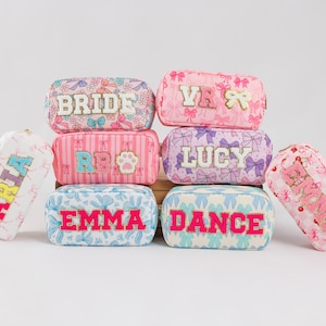 May include: A collection of personalized cosmetic bags in various colors and patterns. Each bag features a name or word in a contrasting color, such as "BRIDE," "LUCY," and "EMMA." The bags are decorated with bows and floral designs.
