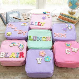 May include: A collection of colorful lunchboxes in various pastel shades, including pink, purple, and mint green. Each lunchbox is personalized with a name or the word "LUNCH" in colorful, textured letters. Decorative patches like rainbows, unicorns, and flowers adorn the boxes.
