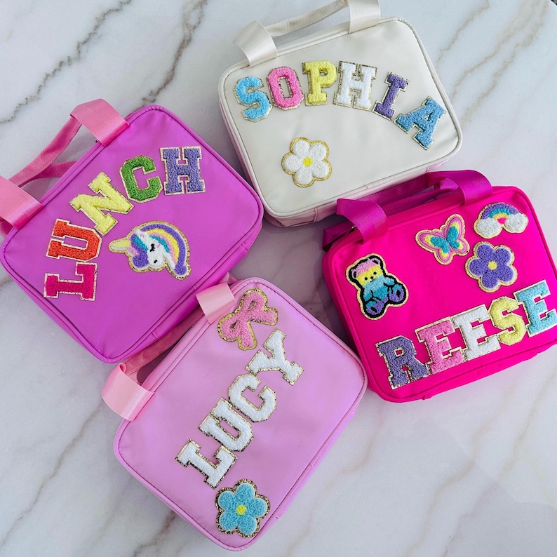 Op de afbeelding: Vier kleurrijke lunchtassen met geborduurde details. E&eacute;n is wit met de naam "SOPHIA", een andere is roze met "LUNCH" en een eenhoorn, een derde is roze met "LUCY", en de laatste is felroze met "REESE" en een teddybeer.