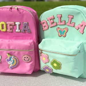 May include: Two colorful backpacks. One is pink with the name "SOFIA" in colorful, textured letters, and patches of a paw print, unicorn, and smiley face. The other is mint green with "BELLA" and a butterfly patch.