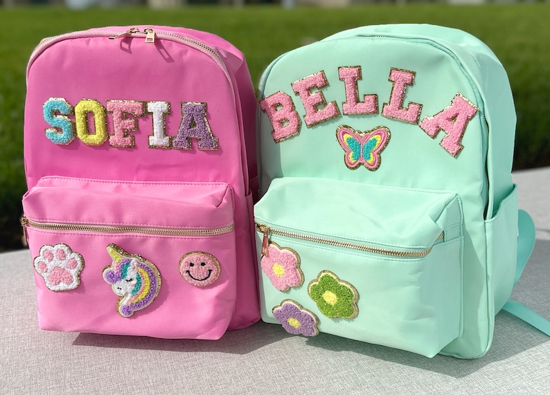 May include: Two colorful backpacks. One is pink with the name "SOFIA" in colorful, textured letters, and patches of a paw print, unicorn, and smiley face. The other is mint green with "BELLA" and a butterfly patch.