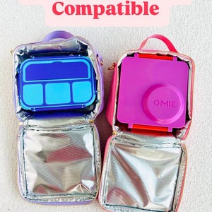 May include: Two open lunch bags, one purple and one pink, each containing a bento box. The purple bag holds a blue and light blue bento box. The pink bag holds a pink bento box. Text at the top reads "Omie & Bento Box Compatible."