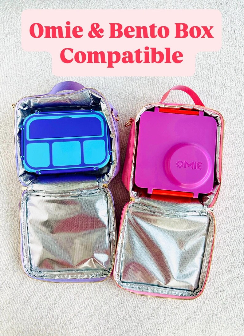 May include: Two open lunch bags, one purple and one pink, each containing a bento box. The purple bag holds a blue and light blue bento box. The pink bag holds a pink bento box. Text at the top reads "Omie & Bento Box Compatible."