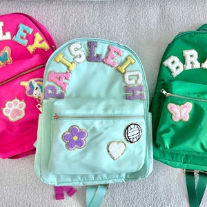 May include: Three personalized backpacks in pink, teal, and green. Each backpack features a name in colorful, embroidered letters. Decorative patches include butterflies, flowers, and a paw print. Zippers and straps are visible.