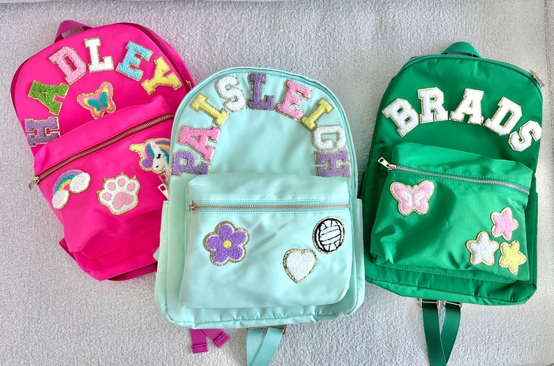 May include: Three personalized backpacks in pink, teal, and green. Each backpack features a name in colorful, embroidered letters. Decorative patches include butterflies, flowers, and a paw print. Zippers and straps are visible.