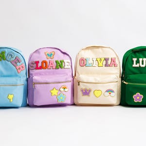 May include: Four colorful backpacks with personalized names: Spencer, Sloane, Olivia, and Lucy. Each backpack features a different color, name patch, and decorative patches like butterflies, stars, and rainbows. They have gold zippers and a front pocket.