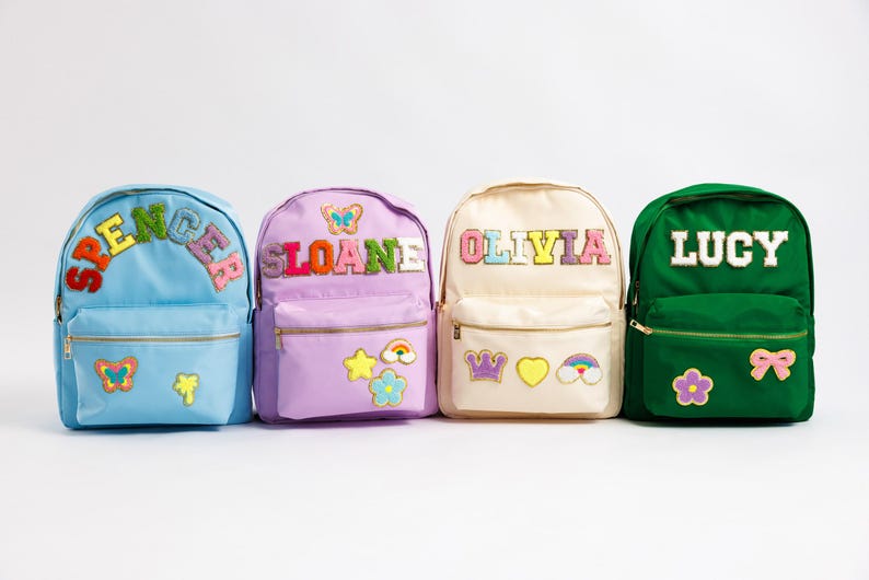 May include: Four colorful backpacks with personalized names: Spencer, Sloane, Olivia, and Lucy. Each backpack features a different color, name patch, and decorative patches like butterflies, stars, and rainbows. They have gold zippers and a front pocket.