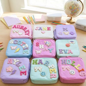 K&ouml;nnte beinhalten: Neun quadratische, pastellfarbene Lunchboxen mit personalisierten Namen in bunten, strukturierten Buchstaben. Jede Box hat dekorative Elemente wie Schmetterlinge, Regenbogen und Smiley-Gesichter. Ein Globus, Bleistifte und Notizb&uuml;cher sind im Hintergrund.