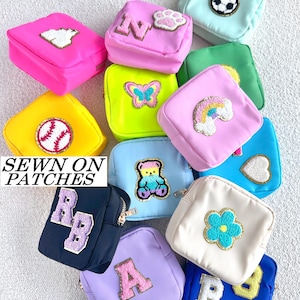 May include: A collection of small, square pouches in various colors, each adorned with sewn-on patches. Patches include letters, a paw print, a rainbow, a butterfly, a soccer ball, a baseball, a teddy bear, a cloud, and a flower. The text "SEWN ON PATCHES" is visible.