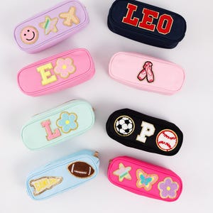 May include: Collection of colorful pencil cases with embroidered patches. Designs include a smiley face, stars, flowers, the name "LEO", ballet shoes, the letters "L" and "E", a soccer ball, a baseball, a football, and a butterfly.