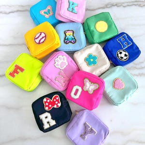 May include: A collection of small, square pouches in various colors, each adorned with a different embroidered patch. Patches include letters, a baseball, a teddy bear, a soccer ball, a paw print, a flower, a butterfly, a heart, and a bow.