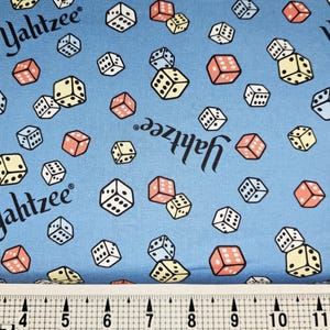 May include: Light blue fabric with a repeating pattern of dice in white, yellow, and coral. The word "Yahtzee" is printed in black script throughout the design. A ruler is at the bottom.